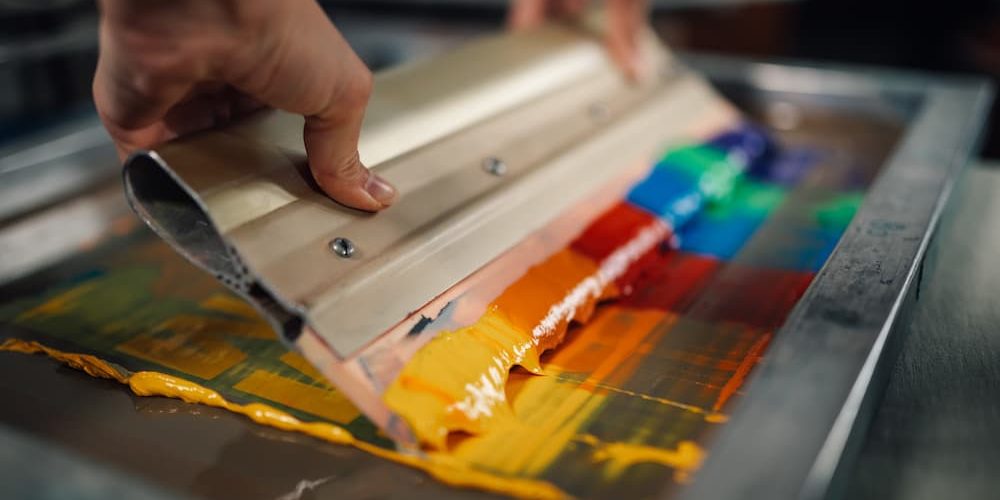 screen printing supplies Close-up of a screen printing squeegee pulling colorful ink across a mesh frame, illustrating high-quality screen printing supplies and custom apparel techniques.