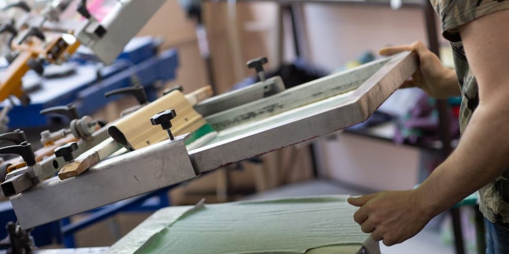 A print shop professional lifting a mesh frame from a garment on a multi-station press, illustrating what is screen printing and how the custom apparel process works.