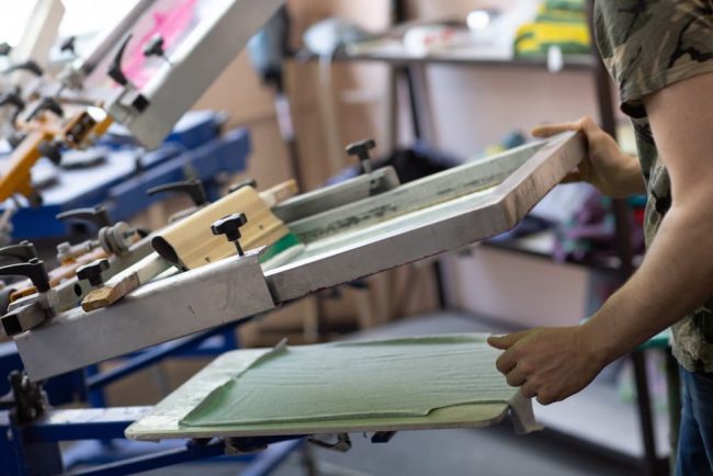 A print shop professional lifting a mesh frame from a garment on a multi-station press, illustrating what is screen printing and how the custom apparel process works.